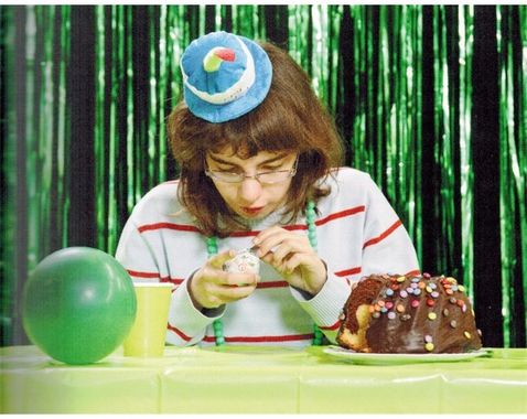 Das Bild zeigt eine junge Frau vor einem grünen Hintergrund. Sie hält etwas in den Händen. Rechts vor ihr ist ein Schokoladenkuchen zu sehen, links von Ihr ein grüner Ball.