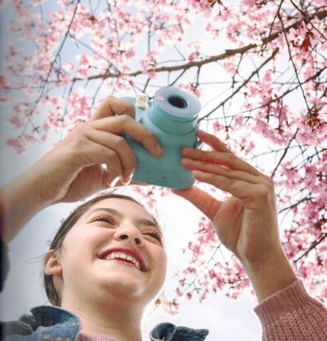 Das Bild zeigt eine lächelnde junge Frau mit einem Fotoapparat unter einem rosa blühenden Baum. Das Bild ist von unten aufgenommen.